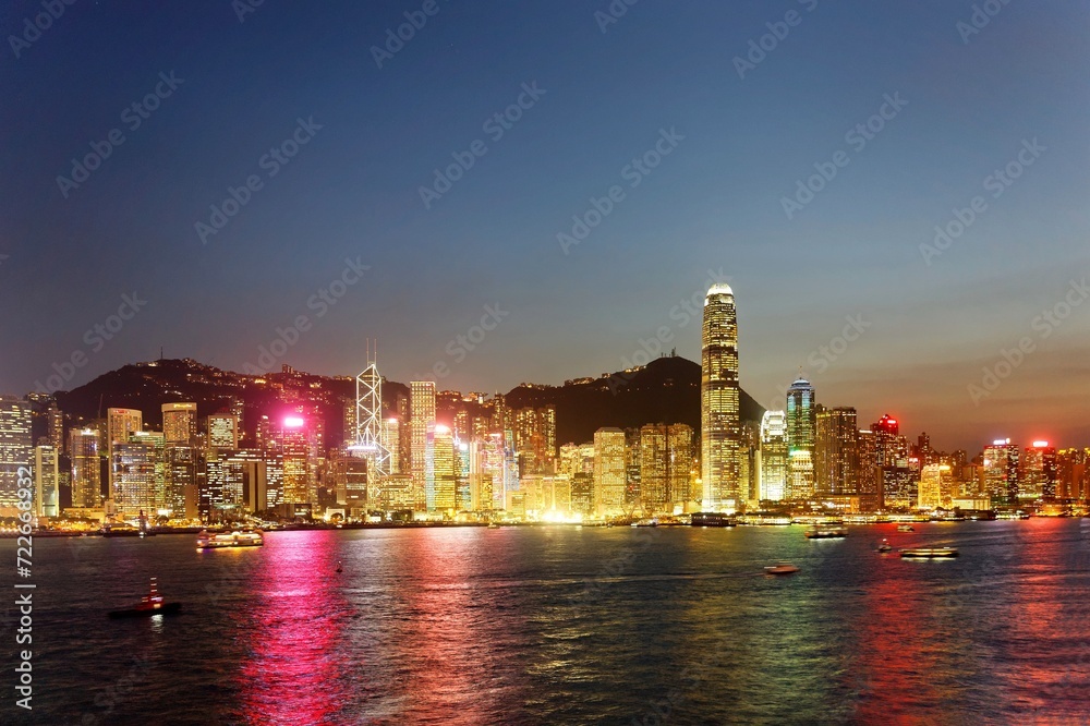 Urban skyline of Hong Kong at dusk with crowded skyscrapers by Victoria Harbor, colorful city lights reflected in the water, Aqua Luna ferry boats crossing the seaport & Victoria Peak under sunset sky