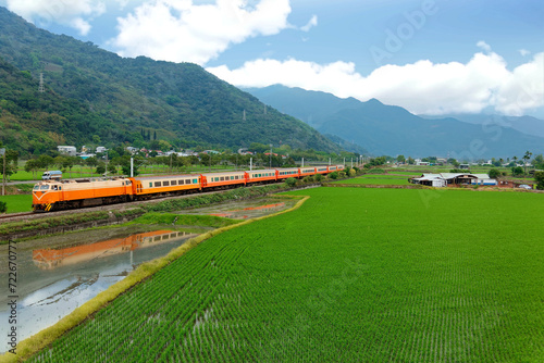 A Chu-Kuang Express train travels between mountains & green fields with reflections in the irrigated rice paddies and newly transplanted seedlings in the farmlands, in Guanshan, Taitung, Taiwan, Asia
