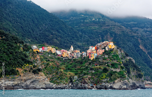 Beautiful scenery of Corniglia viewed from the sea, an amazing village of colorful houses perched on a rocky cliff by the rugged coastline with mountains in background, in Cinque Terre, Italy, Europe