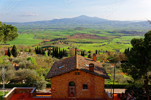 
A brick house on the hillside overlooking the green grassy rolling hills in idyllic Tuscany countryside and Mount Amiata in the background under blue sunny sky, in Old Town Pienza, Val d'Orcia, Italy