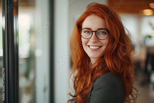 Bright and inviting portrait of a red-haired woman with glasses, smiling confidently in a professional office environment.