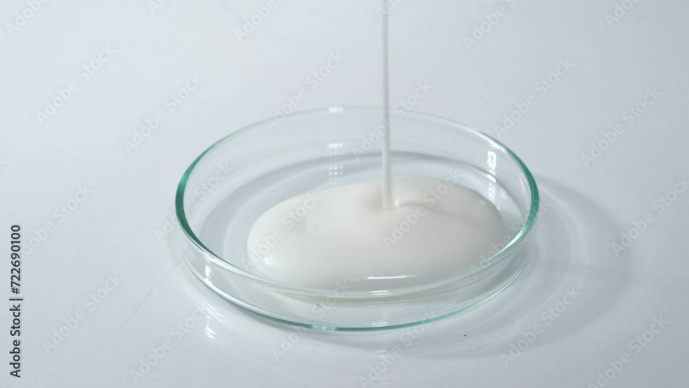 A stream of solid white texture flows down a transparent petri dish against a white background. Laboratory space with new cosmetic research concept. A stream of solid white texture flows down a transparent petri dish against a white background. Laboratory space with new cosmetic research concept.
