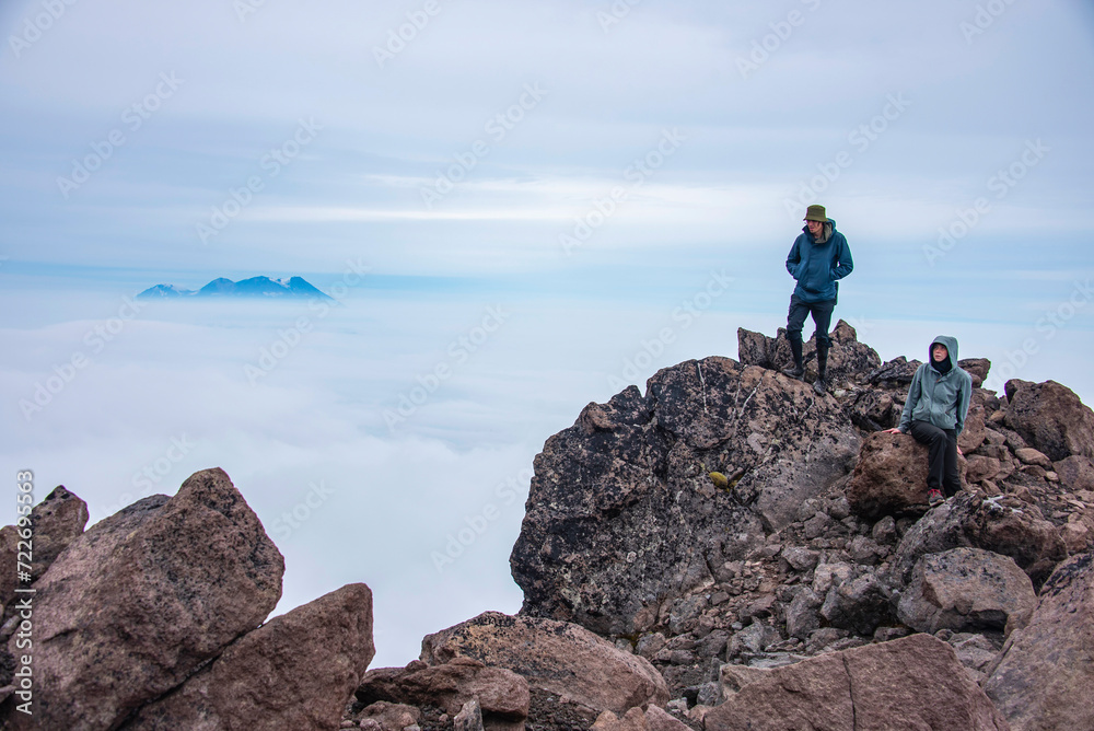 There are man  and woman on the foreground. There is an ocean of clouds after them. There is Zhupanovskiy volcano on the background.