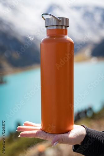 Mockup image of an orange metallic thermos bottle on a female palm in front of the mountain landscape and a lake