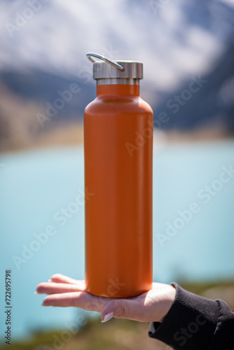 Mockup image of an orange metallic thermos bottle on a female palm in front of the mountain landscape and a lake