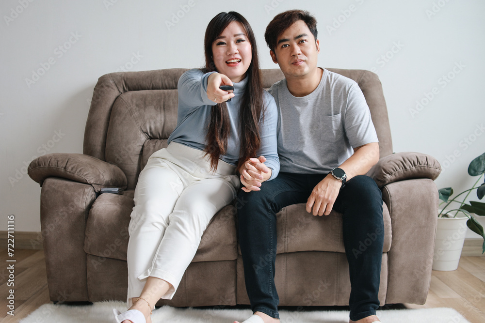 Romantic young Asian couple relaxing on sofa, watching TV and switching channels with remote control