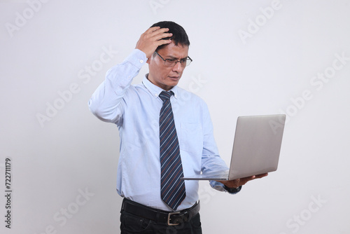 Confused Asian businessman wear glasses and formal office clothes, scratching his head while looking at laptop. 