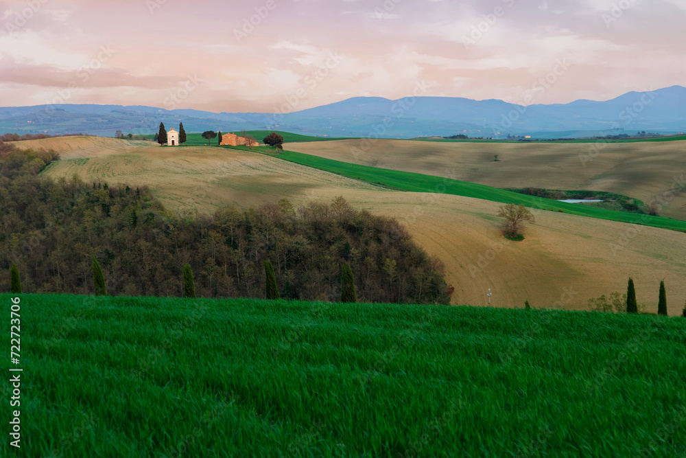 Dusk scenery of a lovely church (The Chapel of Our Lady of Vitaleta ) standing on a grassy hillside with forests in foreground in Pienza, Italy ~ Beautiful sunset scene of rolling hills in Tuscany