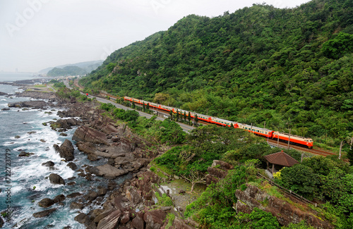 An express train travels on the railway along a coastal highway between a beautiful rocky beach and mountains of green lush forests on a sunny summer day in Beiguan Tidal Park, Toucheng, Yilan, Taiwan