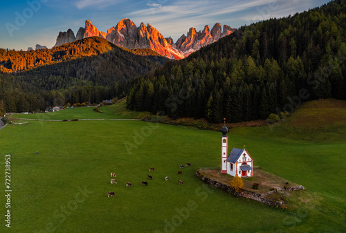 Val Di Funes, Dolomites, Italy - Aerial view of the beautiful St. Johann Church (Chiesetta di San Giovanni in Ranui) at South Tyrol with cows and Italian Dolomites in warm sunset colors at background