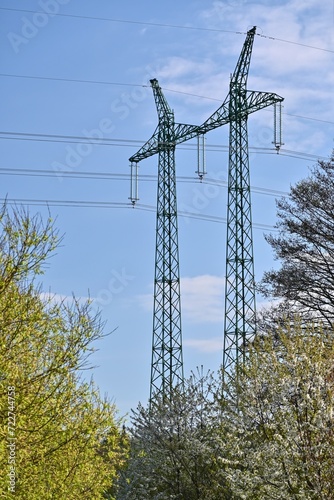 High voltage pole behind blossoming trees and against a cloudy sky background. High voltage transmission line system in the Czech Republic