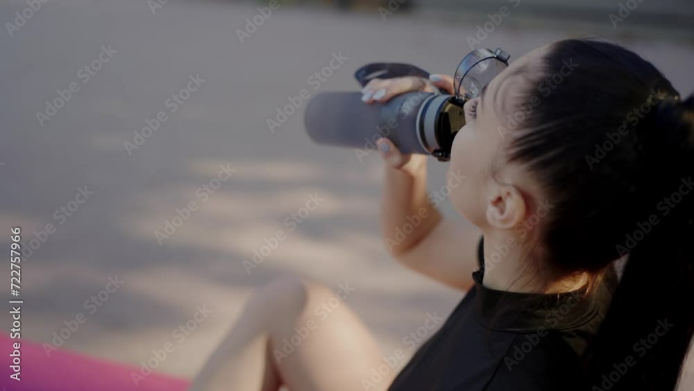 Young woman takes break from exercise to drink water sitting on fitness mat. Young sportswoman prefers doing sports in fresh air on ground in warm summer time. Active lifestyle Young woman takes break from exercise to drink water sitting on fitness mat. Young sportswoman prefers doing sports in fresh air on ground in warm summer time. Active lifestyle