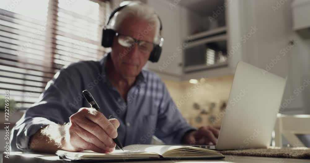 Home laptop, notebook and old man writing business information, schedule or summary of online report in kitchen. Headphones, journal and senior person doing research, remote work and planning agenda