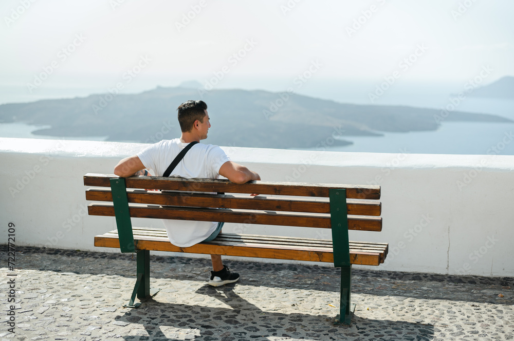 Back view of young man sitting on bench looking at Santorini coastline