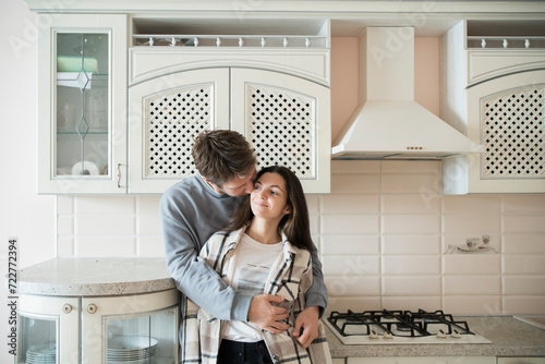 A couple stands close together in an affectionate embrace, exchanging loving looks in the comfort of their sunlit kitchen. © Mihail