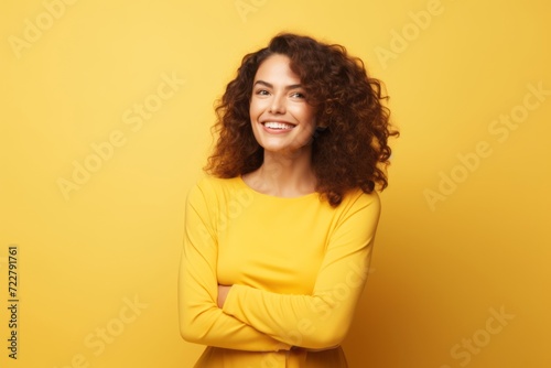 Portrait of a smiling young woman with curly hair standing with crossed arms over yellow background