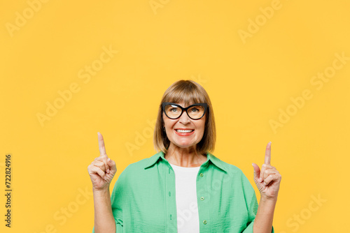 Elderly blonde woman 50s year old wear green shirt glasses casual clothes point index finger overhead on area indicate on area copy space mock up isolated on plain yellow background Lifestyle concept photo