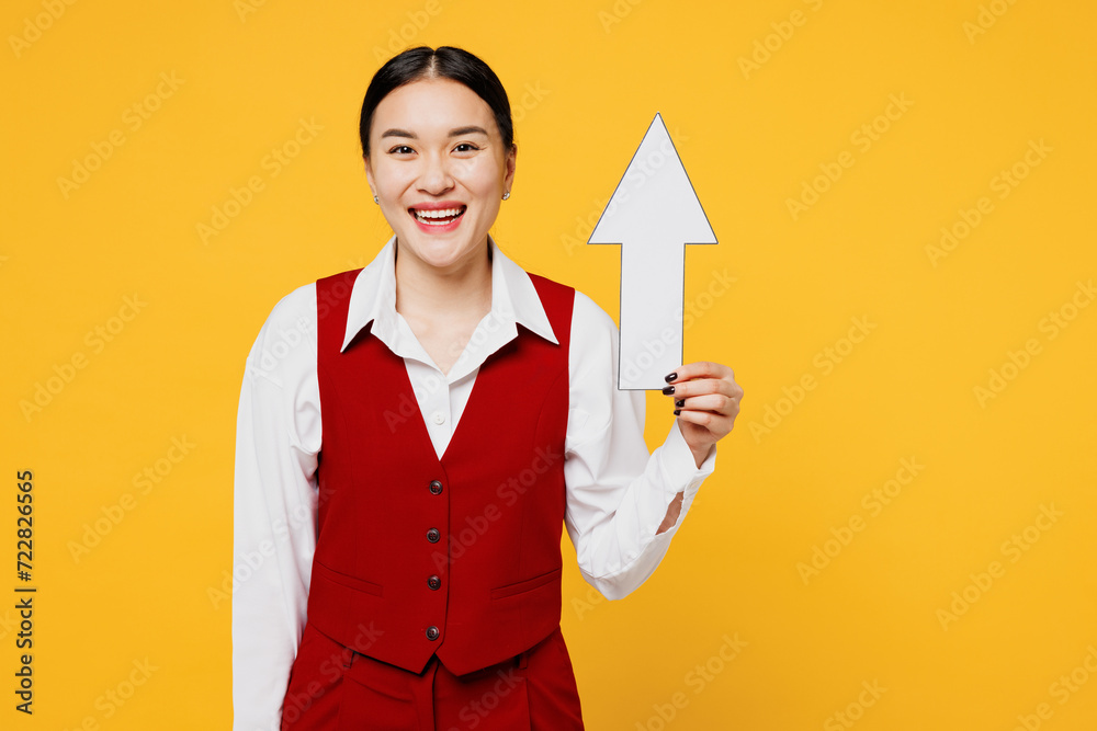 Young smiling happy corporate lawyer employee business woman of Asian ethnicity wear formal red vest shirt work at office holding up arrow isolated on plain yellow background studio. Career concept. Young smiling happy corporate lawyer employee business woman of Asian ethnicity wear formal red vest shirt work at office holding up arrow isolated on plain yellow background studio. Career concept.