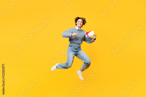 Full body young woman she wears grey knitted sweater shirt casual clothes jump high hold point on gift certificate coupon voucher card for store isolated on plain yellow background. Lifestyle concept.