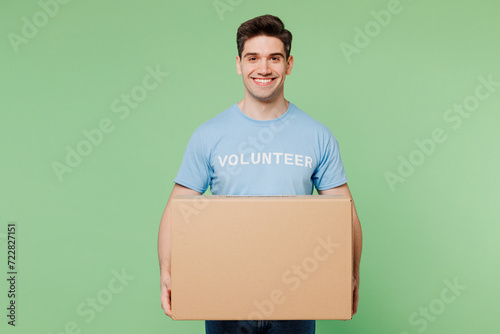 Young smiling man wears blue t-shirt white title volunteer hold in hand blank cardboard box isolated on plain pastel green background. Voluntary free team work assistance help charity grace concept. photo