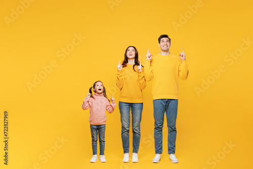 Full body young happy parents mom dad with child kid girl 7-8 years old wear pink sweater casual clothes point index finger overhead on area isolated on plain yellow background. Family day concept. photo