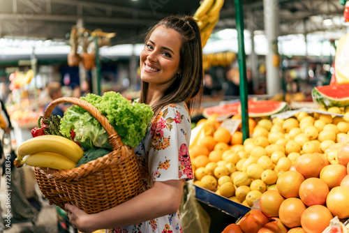 A portrait of a smiling Caucasian girl holding a basket filled with fruits and vegetables she bought at the market.