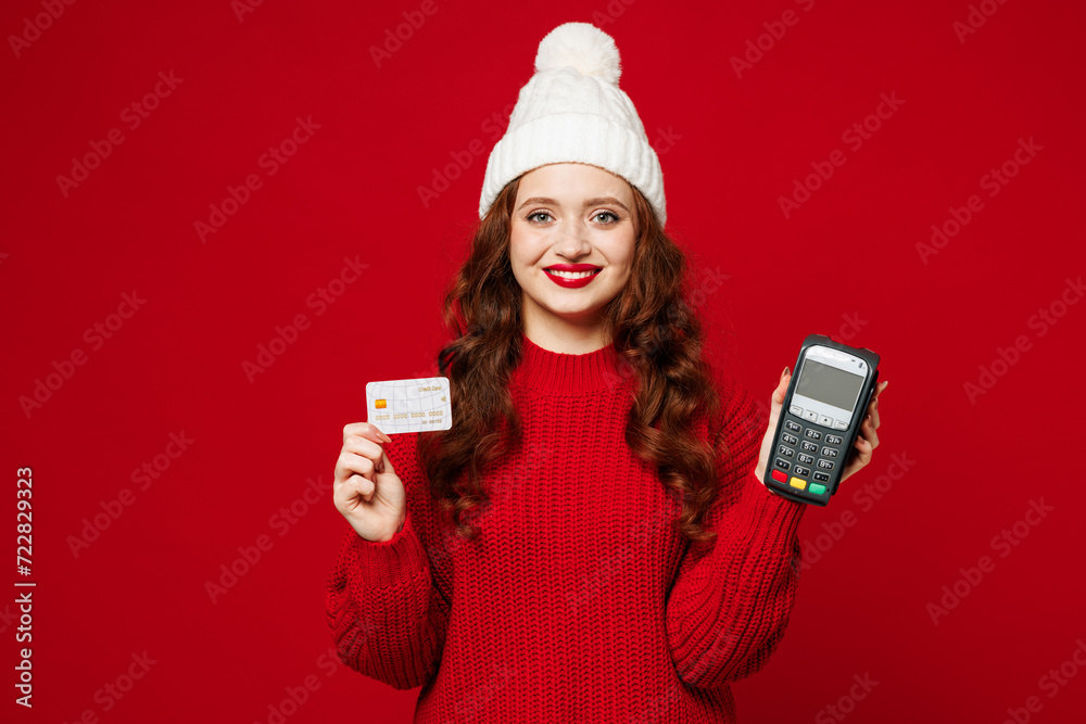 Young woman wears knitted sweater white hat casual clothes hold wireless modern bank payment terminal to process acquire credit card isolated on plain red color background studio. Lifestyle concept.