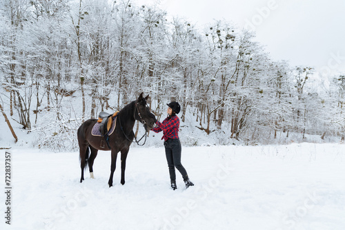 A beautiful brunette girl in a plaid black-and-white shirt walks with a big black horse in a snowy park. A woman in a helmet for riding walks in a snowy forest with a thoroughbred horse