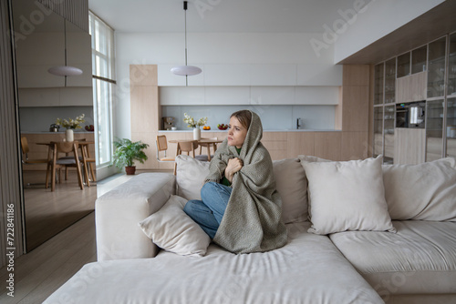 Sad tired woman sits on couch at home wrapped in plaid in living room. Blue mood of depressed girl boring. Nervous female feeling frustration hiding from society in loneliness solitude. Life problems. photo