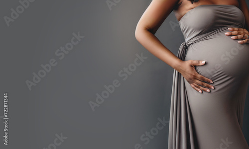 A pregnant African American woman in a gray dress holds her stomach on an acer background. Copy space