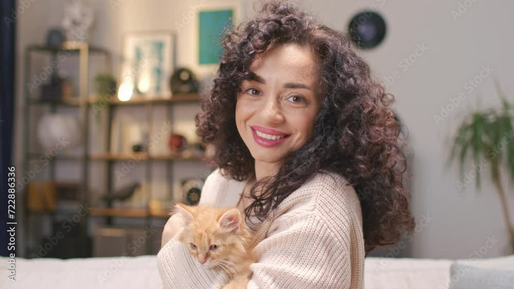 Zooming in on young Caucasian woman hugging little red cat and joyfully smiling on camera. Curly female owner petting her lovely kitten in cozy room at home. Concept of love to animals.