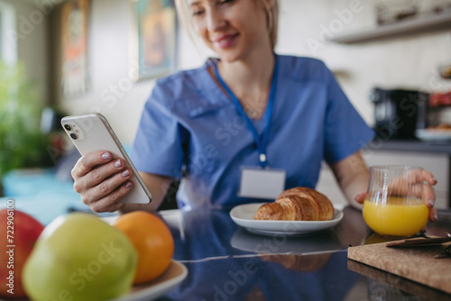 Female nurse or doctor getting ready for work in the morning, scrolling on smartphone while eating breakfast. Work-life balance for healthcare worker. photo