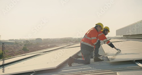 Two engineer workers in safety gear install solar panels on a rooftop, with a suburban landscape stretching out in the distance