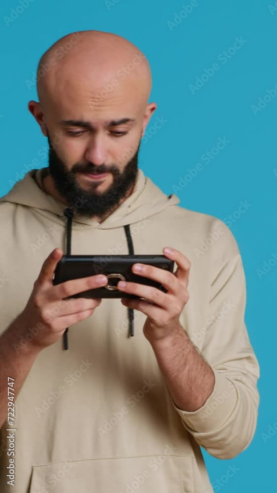 Vertical Video Joyful muslim man playing mobile video games in studio, enjoying online competition using smartphone app. Arabic gamer having fun with roleplaying challenge contest. Camera 1. Vertical Video Joyful muslim man playing mobile video games in studio, enjoying online competition using smartphone app. Arabic gamer having fun with roleplaying challenge contest. Camera 1.