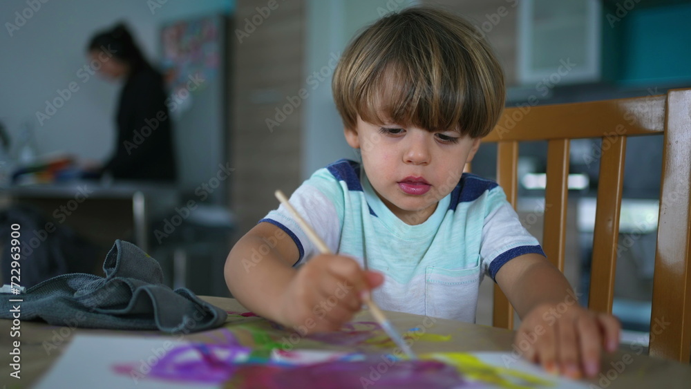 Artistic child plays with watercolor paint at home while mother does chores in background. Creative little boy painting with brush