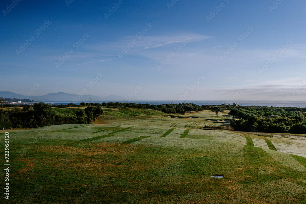 Sotogrande, Spain - January, 23, 2024 - Elevated view of a golf course with mowed fairways leading towards the sea, with mountains and a clear sky in the background.