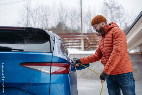 Man charging electric car during cold snowy day. Side view of hansome man putting charger in charging port during winter, cold weather. Charging and driving electric vehicles during winter season.
