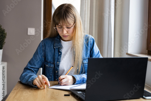 Young woman freelancer in casual clothes sitting at table using laptop working or studying online at home