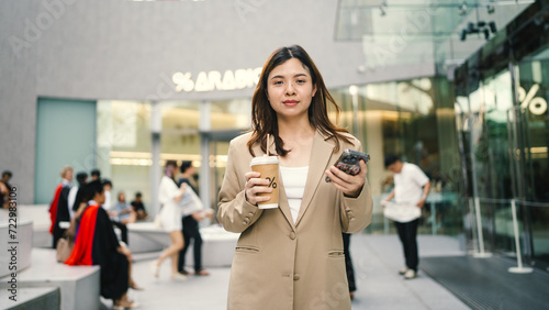 Young asian elegant business woman leader wearing suit standing in modern city building using cell phone platform applications hold coffee drink