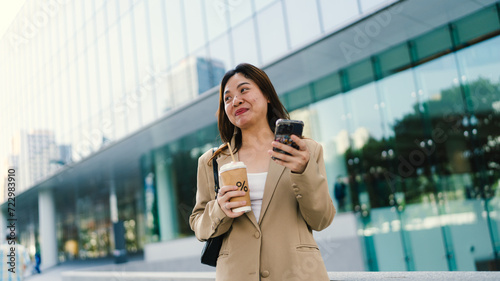 Young asian elegant business woman leader wearing suit standing in modern city building using cell phone platform applications hold coffee drink