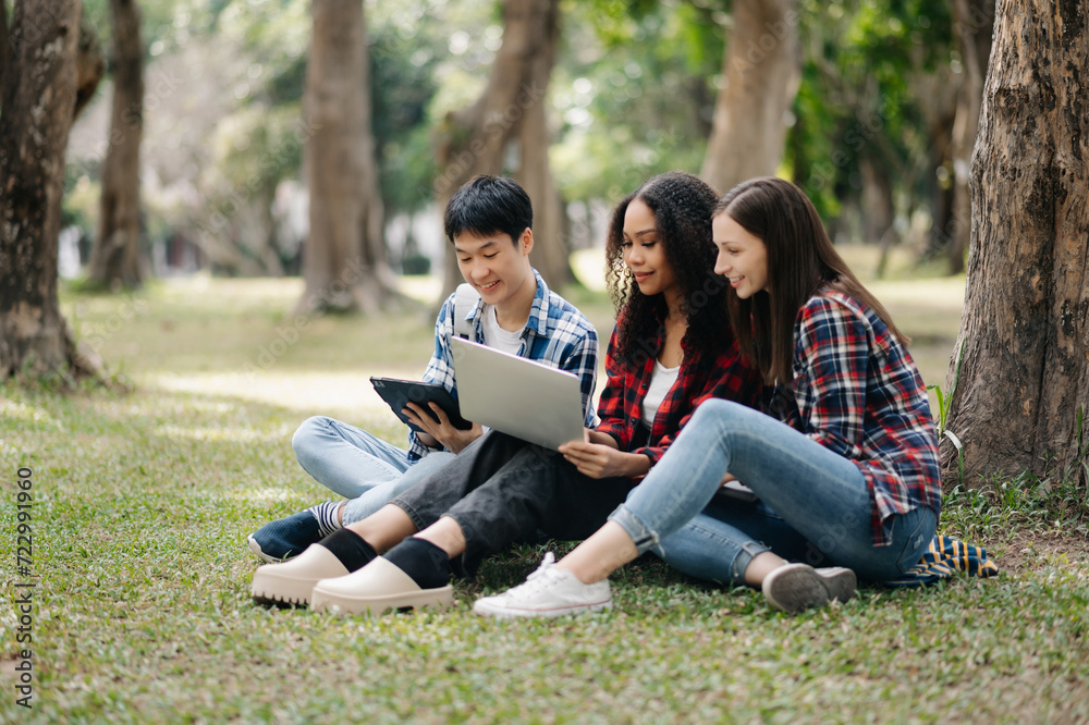 Three young college students is reading a book while relaxing sitting on grass in a campus park with her friends. Education concept Three young college students is reading a book while relaxing sitting on grass in a campus park with her friends. Education concept