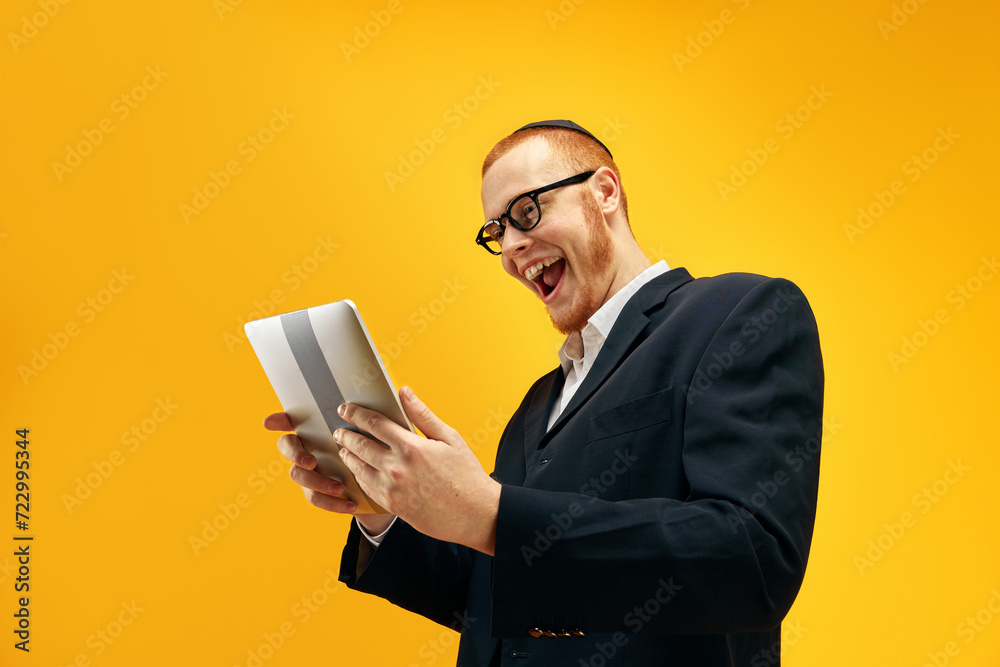 Emotional Jewish young man in glasses, looking on tablet with excitement, reading news against yellow studio background. Concept of Purim holiday, Jewish traditions, history and culture Emotional Jewish young man in glasses, looking on tablet with excitement, reading news against yellow studio background. Concept of Purim holiday, Jewish traditions, history and culture
