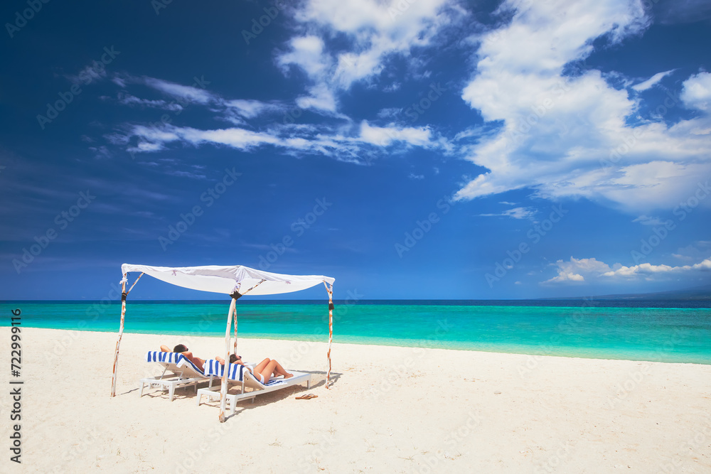 Happy couple relaxing on tropical beach on sun loungers under white umbrella against backdrop of beautiful sky and islands during summer vacation. Beach holiday concept