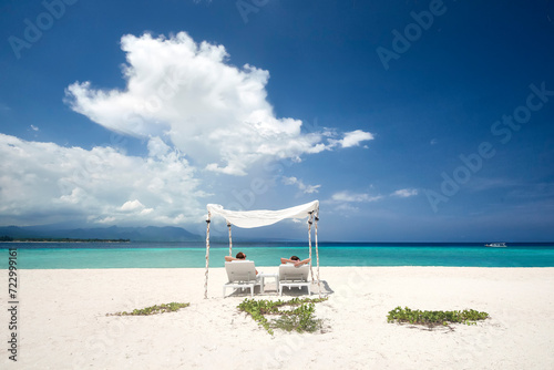 Happy couple relaxing on tropical beach on sun loungers under white umbrella against backdrop of beautiful sky and islands during summer vacation. Beach holiday and travel concept © soft_light
