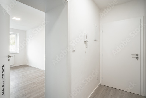 An empty room with white walls  a closed entrance door  and an opening leading to a naturally lit room. There is a light switch and intercom by the door.