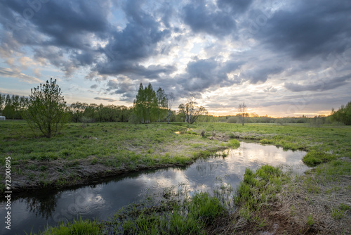 The sunset sky is reflected in the stream, a green field in the spring at sunset