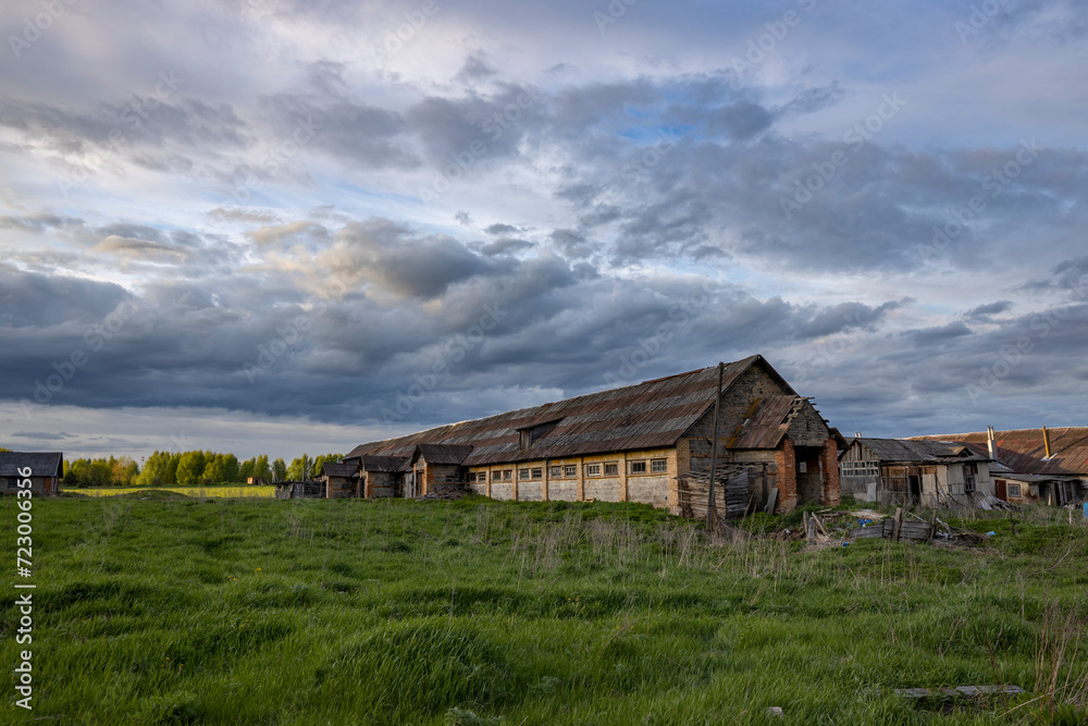 An old abandoned cowshed against the backdrop of a spring evening landscape with clouds and green grass