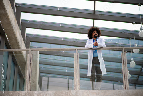 Portrait of doctor standing in hospital, leaning on glass railing, looking at camera. © Halfpoint