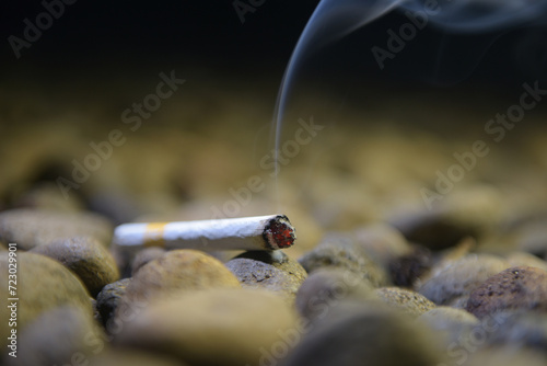 a cigarette with burning embers and ash at the end that emits white smoke between the rocks