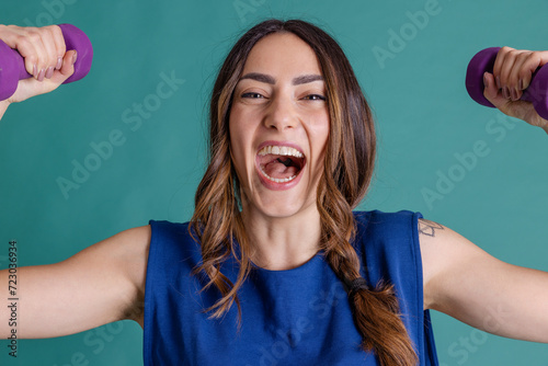 Cheerful young woman holding dumbbells laughing against blue background photo
